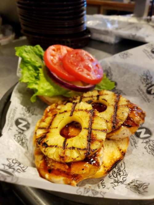 Grilled chicken sandwich topped with pineapple rings, lettuce, tomato, and onion on a paper-lined plate.
