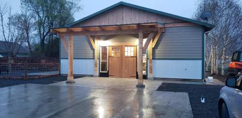 A modern house with a wooden porch, large front door, and a gravel driveway, illuminated in the evening light.