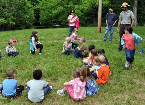 A group of children sitting on grass, listening to an adult in a ranger hat, with adults observing in the background.
