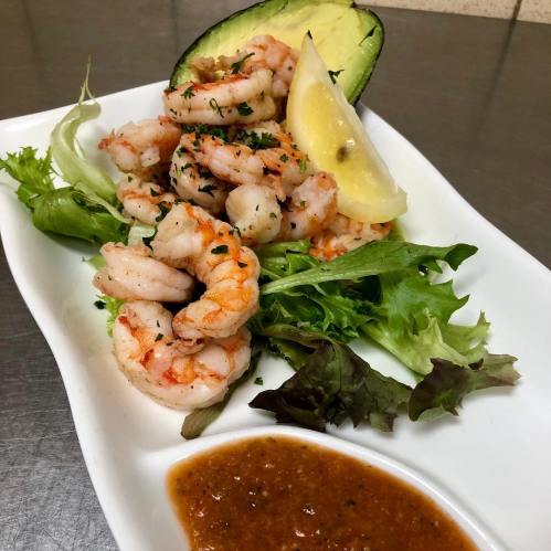 A plate of shrimp garnished with avocado, lemon, and mixed greens, served with a side of sauce.