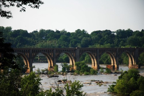 A historic stone bridge with multiple arches spans a river, surrounded by lush greenery and trees.
