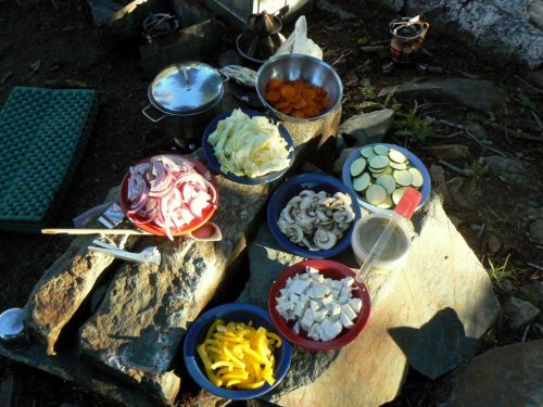 A variety of chopped vegetables and tofu arranged in bowls on rocks, with cooking pots and camping gear nearby.