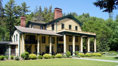 A large, historic yellow house with green accents, featuring columns and a manicured lawn surrounded by trees.