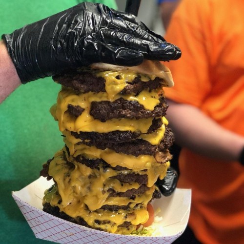 A towering burger stacked with multiple beef patties and melted cheese, held by a gloved hand.