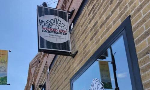 Sign for "Wicked Hen Farmhouse" featuring a logo and text, mounted on a brick wall.