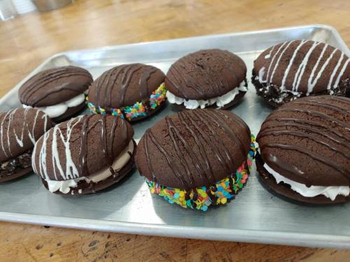 A tray of chocolate whoopie pies filled with cream, some decorated with colorful sprinkles and drizzled chocolate.