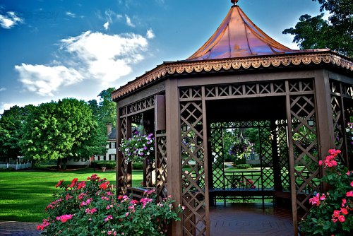A wooden gazebo with a copper roof surrounded by vibrant flowers and lush green grass under a blue sky.