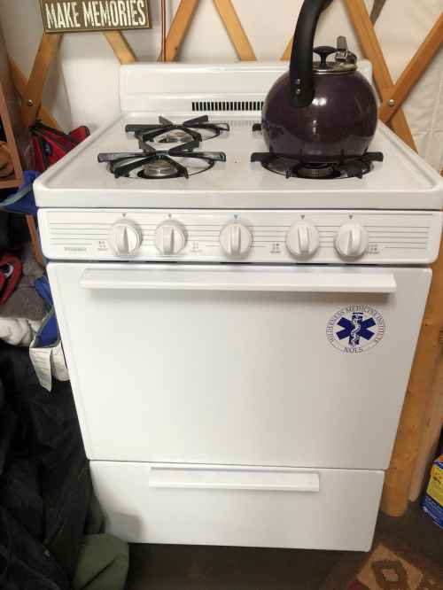 A white gas stove with four burners and a kettle on top, located in a cozy, cluttered space.