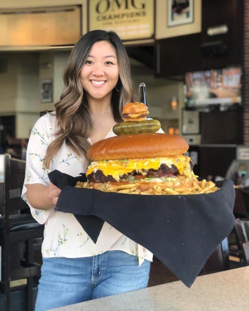 A woman holds a giant burger topped with pickles and a small burger, surrounded by fries, in a restaurant setting.