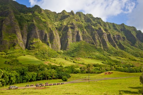 Lush green mountains rise dramatically under a blue sky, with a line of vehicles parked in a scenic valley below.