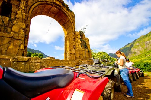 A woman prepares an ATV near ancient ruins, surrounded by lush green mountains and a blue sky.