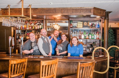 A group of five staff members stands behind a bar, smiling and holding drinks, with shelves of liquor in the background.