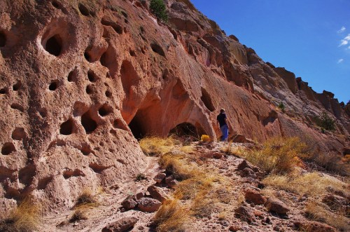 A person stands near a rocky cliff with numerous holes, surrounded by dry grass and a clear blue sky.