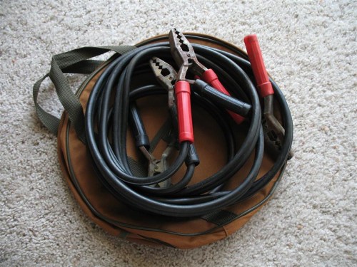 A set of jumper cables with red and black clamps, resting on a brown carrying bag on a carpeted surface.