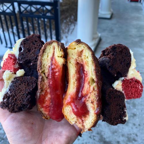 A hand holding a split pastry filled with strawberry jam, surrounded by pieces of chocolate and red velvet cake.