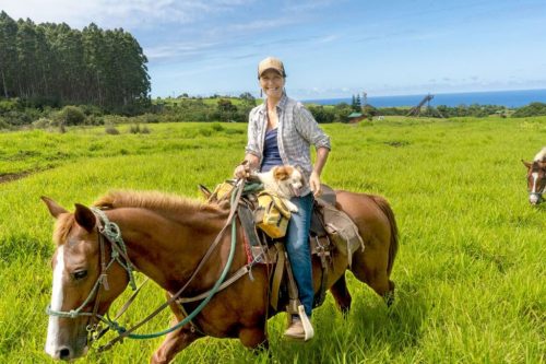 A woman rides a horse in a green field, holding a dog, with trees and ocean in the background.