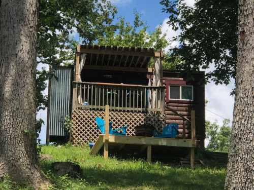 A rustic cabin with a porch, surrounded by trees and grass, featuring blue chairs on a wooden deck.