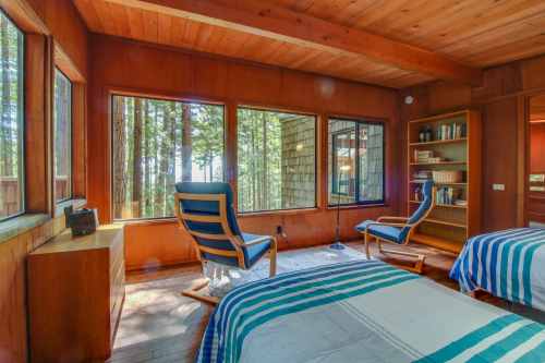 Cozy bedroom with wooden walls, large windows, and striped bedding, surrounded by trees. A chair and bookshelf are visible.