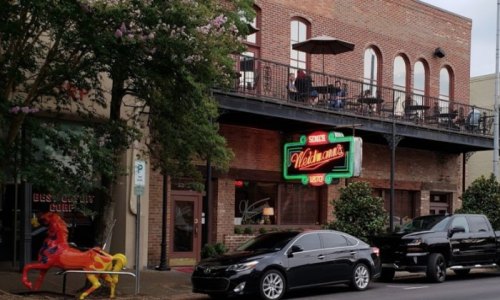 A street view of a brick building with a neon sign, outdoor seating, and a colorful horse sculpture in front.