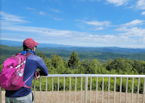 A person in a cap and backpack gazes at a scenic mountain view under a blue sky with scattered clouds.