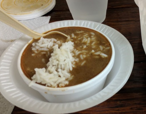 A bowl of brown stew with rice on top, served with a wooden spoon, on a paper plate.