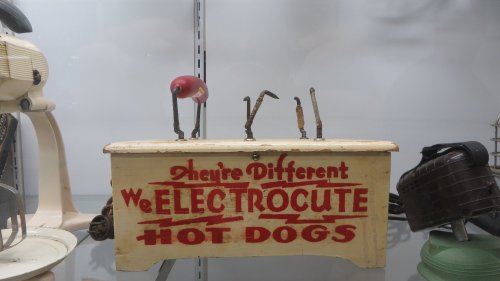 A vintage hot dog cooker with the text "We ELECTROCUTE HOT DOGS" displayed in a glass case.