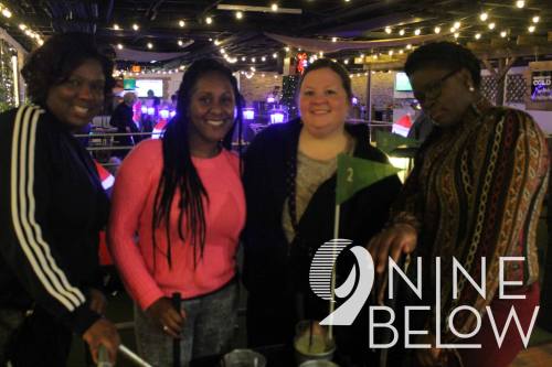 Four friends smiling and posing together at a lively indoor venue with string lights and a bar in the background.