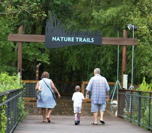 A family walks towards a "Nature Trails" sign on a wooden path surrounded by greenery.