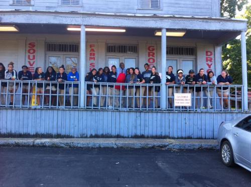 A group of people stands on a porch in front of a rustic building with signs for a grocery store and farm.