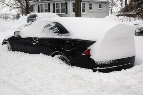 A black car partially buried in deep snow, with only the top visible, in a residential area.