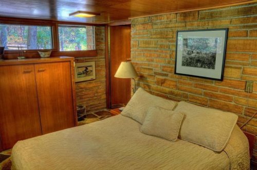 Cozy bedroom with a stone wall, wooden furniture, a lamp, and a framed photo, featuring a warm, inviting atmosphere.