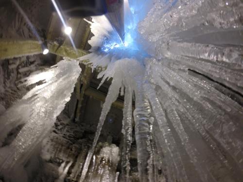 Icicles hang from a ceiling, illuminated by blue light, creating a striking winter scene.