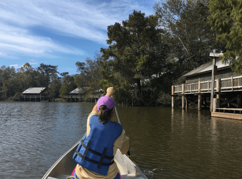 A person paddles a canoe on a calm river, surrounded by trees and wooden cabins on stilts.
