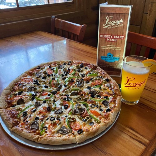 A large pizza topped with vegetables and olives, alongside a drink and a menu at Loopy's Saloon & Grill.