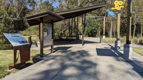 A park shelter with informational signage, surrounded by trees and a pathway, featuring colorful decorations.