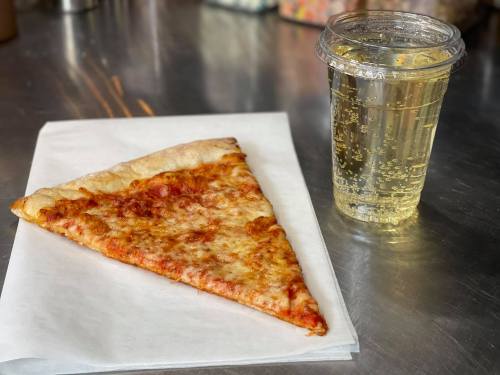 A slice of cheese pizza on a white napkin next to a clear cup of sparkling beverage.