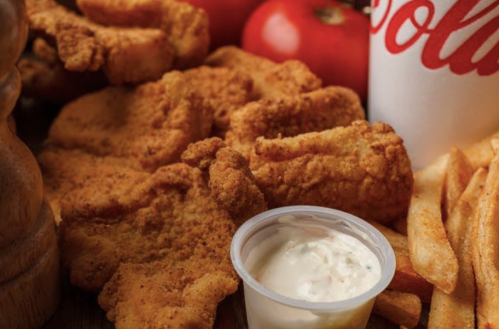 A close-up of crispy fried chicken tenders, fries, a cup of dipping sauce, and fresh tomatoes on a wooden surface.