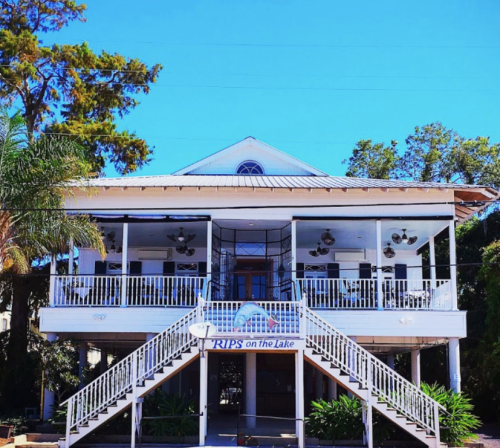 A two-story building with a white exterior, large porch, and fans, surrounded by trees and blue sky.