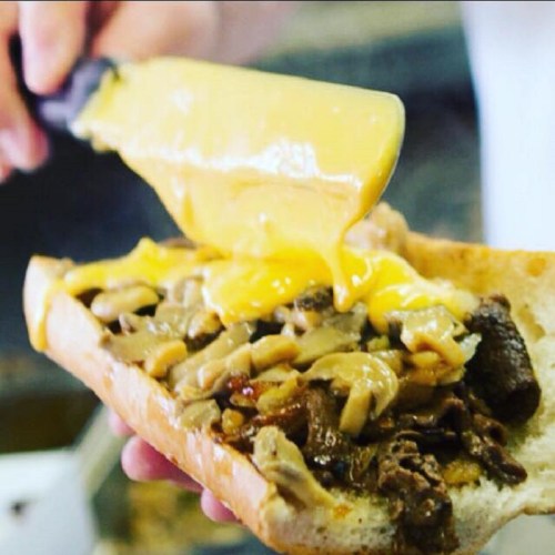 A hand holds a sandwich filled with beef and mushrooms, topped with melted cheese being poured over it.