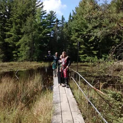 Three individuals in medieval attire stand on a wooden bridge over a marshy area, surrounded by trees and greenery.