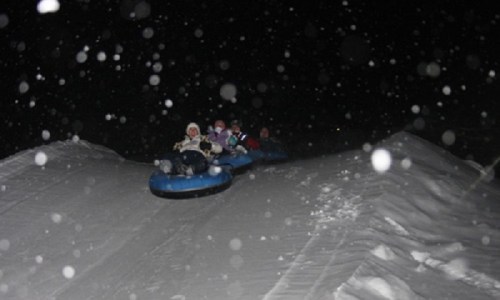 A group of people tubing down a snowy hill at night, with snowflakes falling around them.