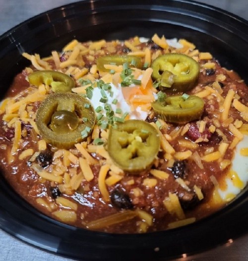 A close-up of chili topped with cheese, jalapeños, and a dollop of sour cream in a black bowl.