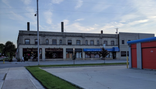 A brick building with shops, including a bookstore and an Asian market, beside a storage facility on a quiet street.