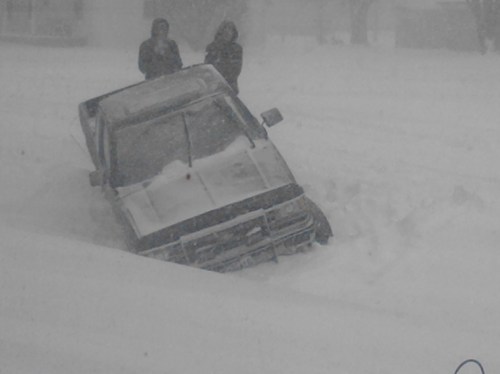 A truck stuck in deep snow with two people standing nearby during a heavy snowstorm.