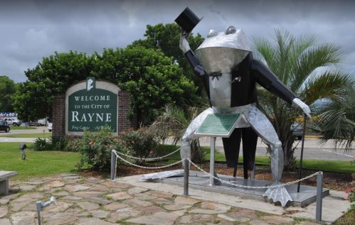 A large metallic frog statue in a tuxedo stands next to a welcome sign for the city of Rayne, Louisiana.