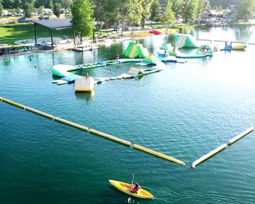 Aerial view of a lake with inflatable water park features and a person kayaking nearby. Trees and a pavilion in the background.