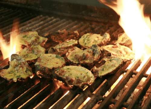 Grilled oysters topped with cheese and herbs, sizzling over an open flame on a barbecue grill.