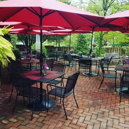 Outdoor dining area with red umbrellas, black tables, and chairs, surrounded by greenery and potted plants.