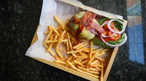 A wooden tray with a cheeseburger topped with bacon, lettuce, tomato, and onion, alongside a serving of crispy fries.
