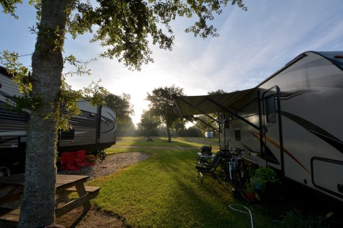A serene RV park scene with two RVs, a picnic table, and bicycles under a sunny sky and trees.